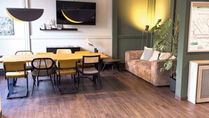 A modern living room with a dining area, featuring a wooden floor, a sofa, a TV mounted on the wall, and a large window. - Coffee Fellows Hotel Frankfurt-Langen (Langen)