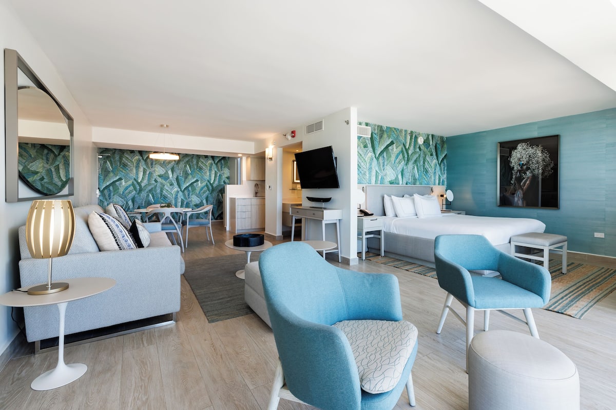 A modern hotel room with a sofa, two armchairs, a bed, a TV, and a dining area.
