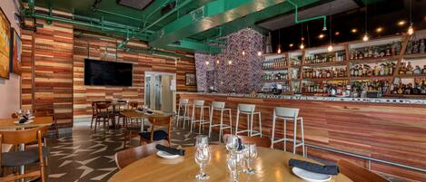 A restaurant with wooden panel walls, a bar area with shelves of bottles, and tables set with glasses and napkins.