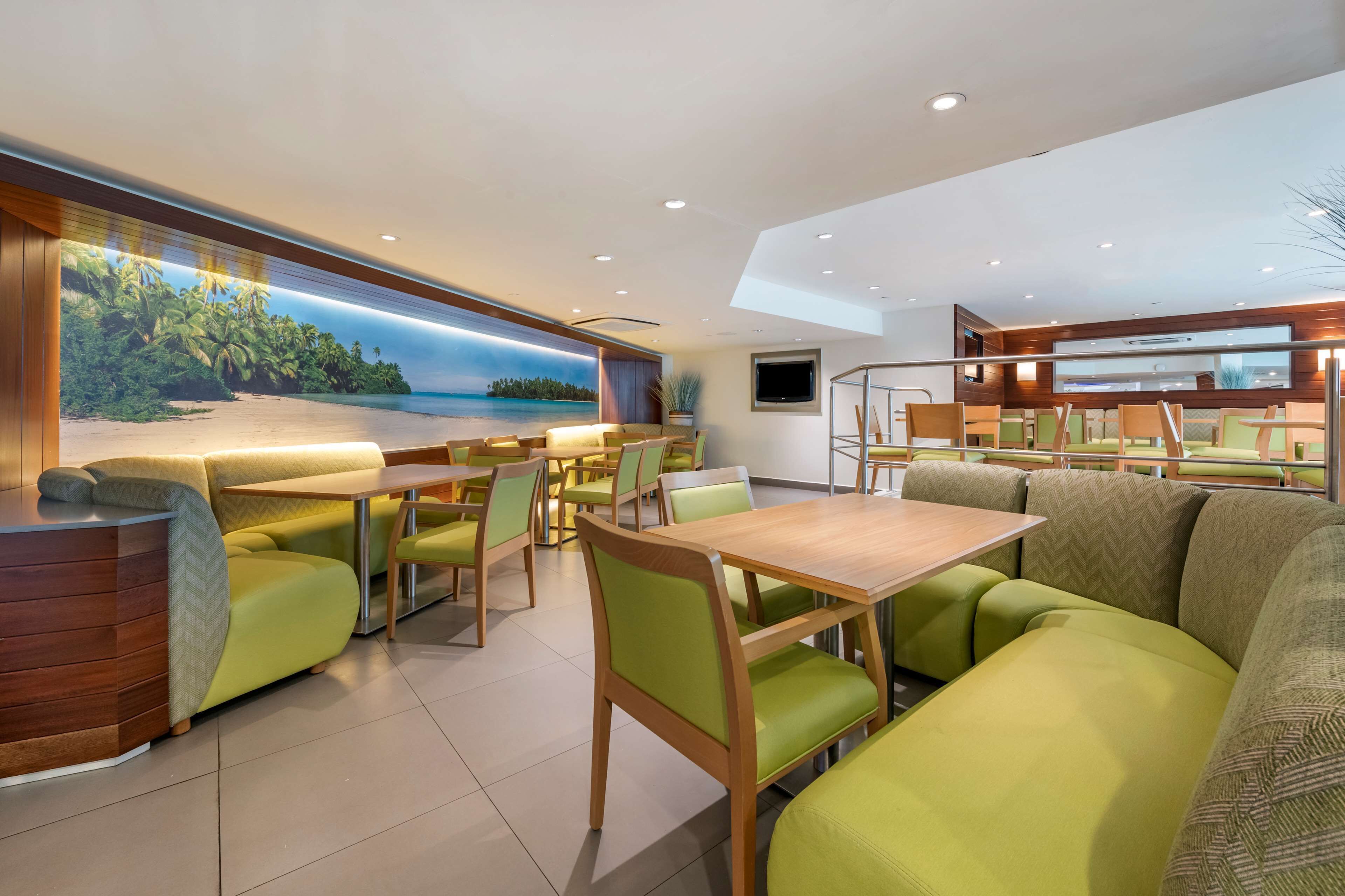 Un restaurante moderno con asientos tapizados en verde, mesas de madera y un gran mural con una escena de playa.