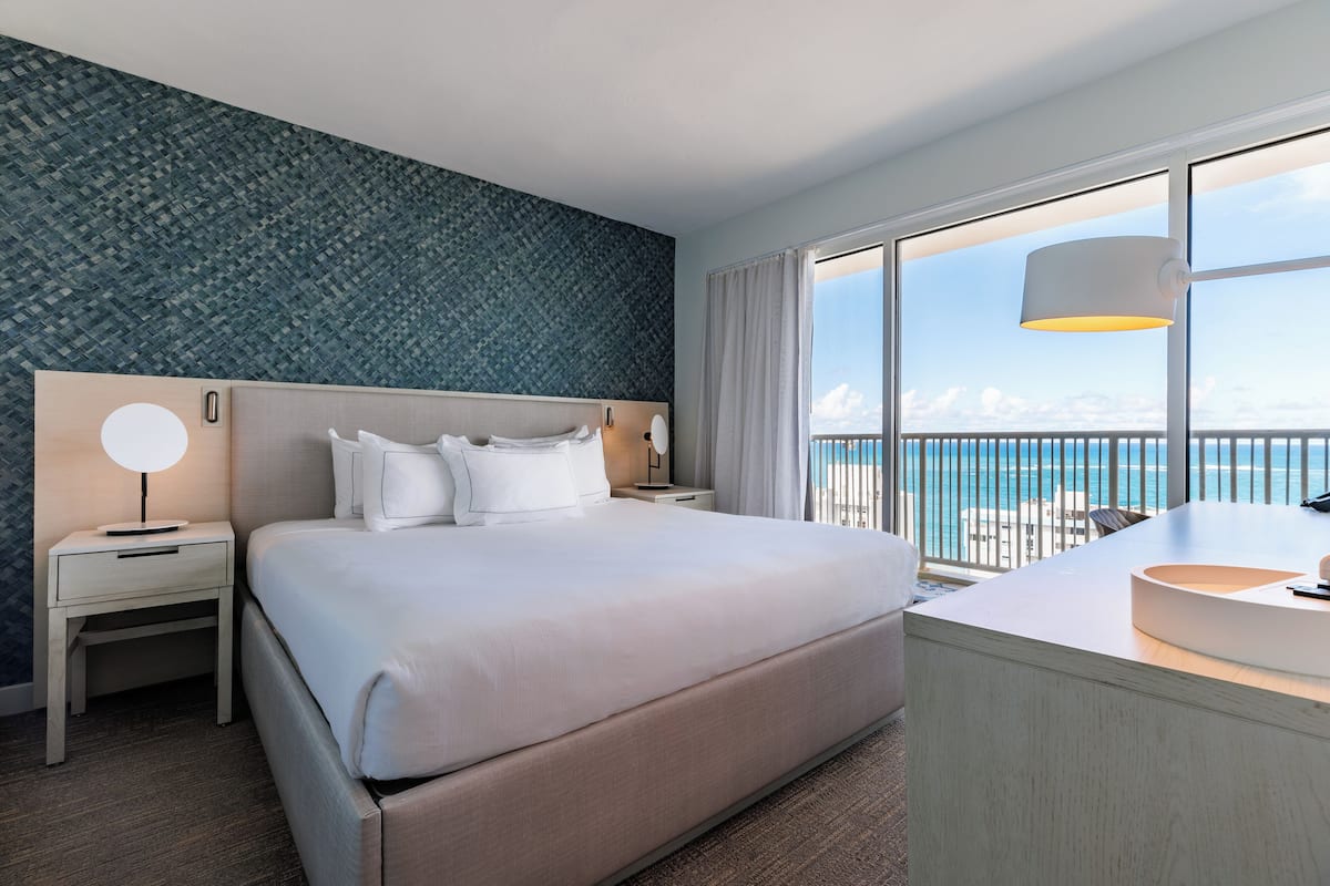 A hotel room with a large bed, a bedside table with a lamp, a bathroom with a sink, and a view of the ocean through large windows.