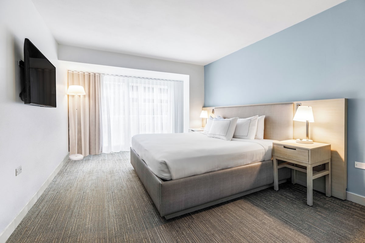 A hotel room with a bed, bedside tables, a TV mounted on the wall, and a window with curtains.