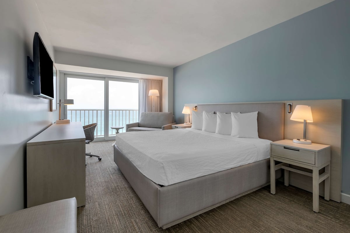 A hotel room with a large bed, a desk, a chair, a TV, and a balcony with a view.