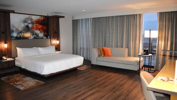Ein modernes Hotelzimmer mit einem großen Bett, einer Couch, einem Schreibtisch und einem großen Fenster mit Vorhängen.