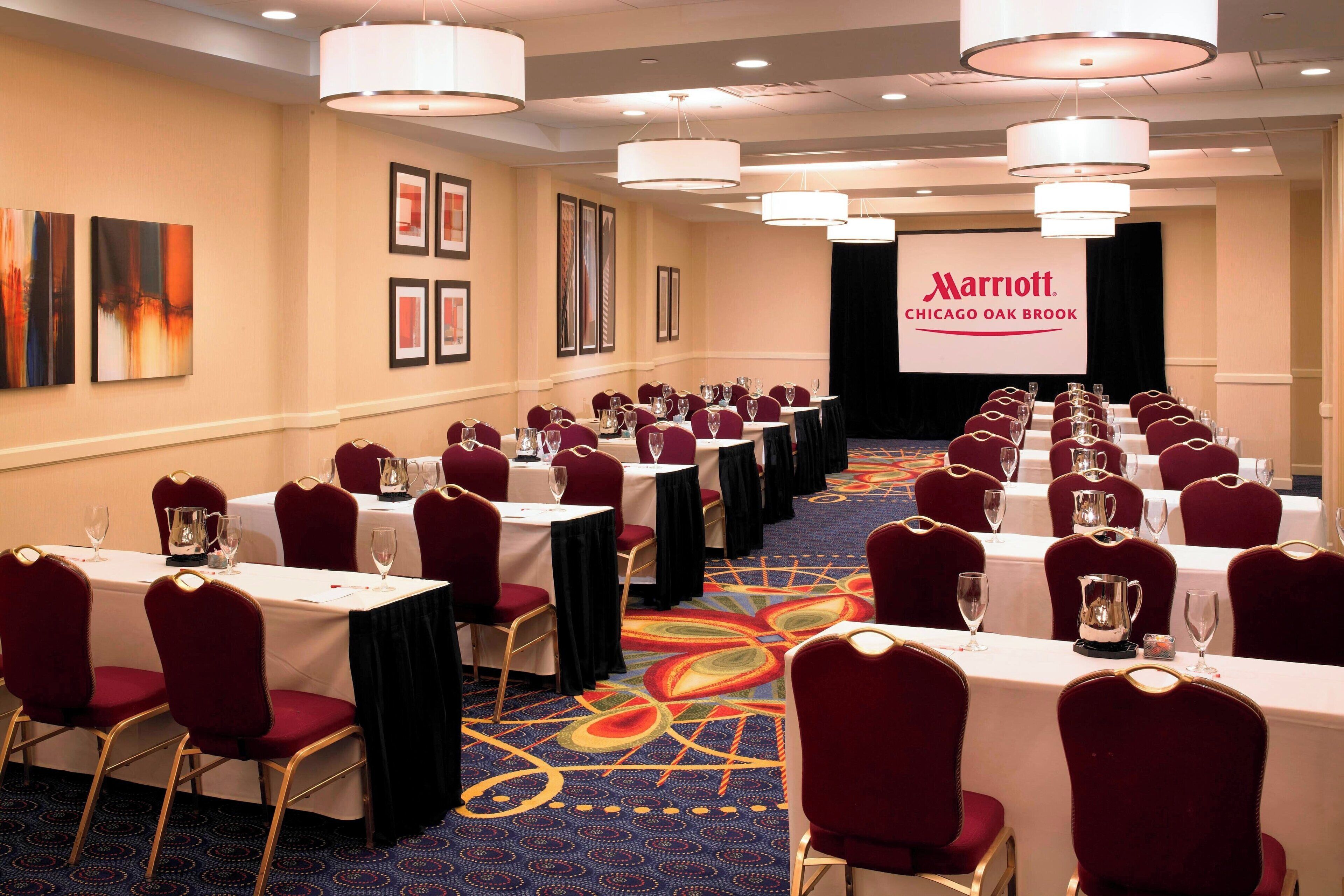 Une salle de conférence avec des tables disposées pour une réunion, des chaises rouges et un grand écran affichant le logo de Marriott.