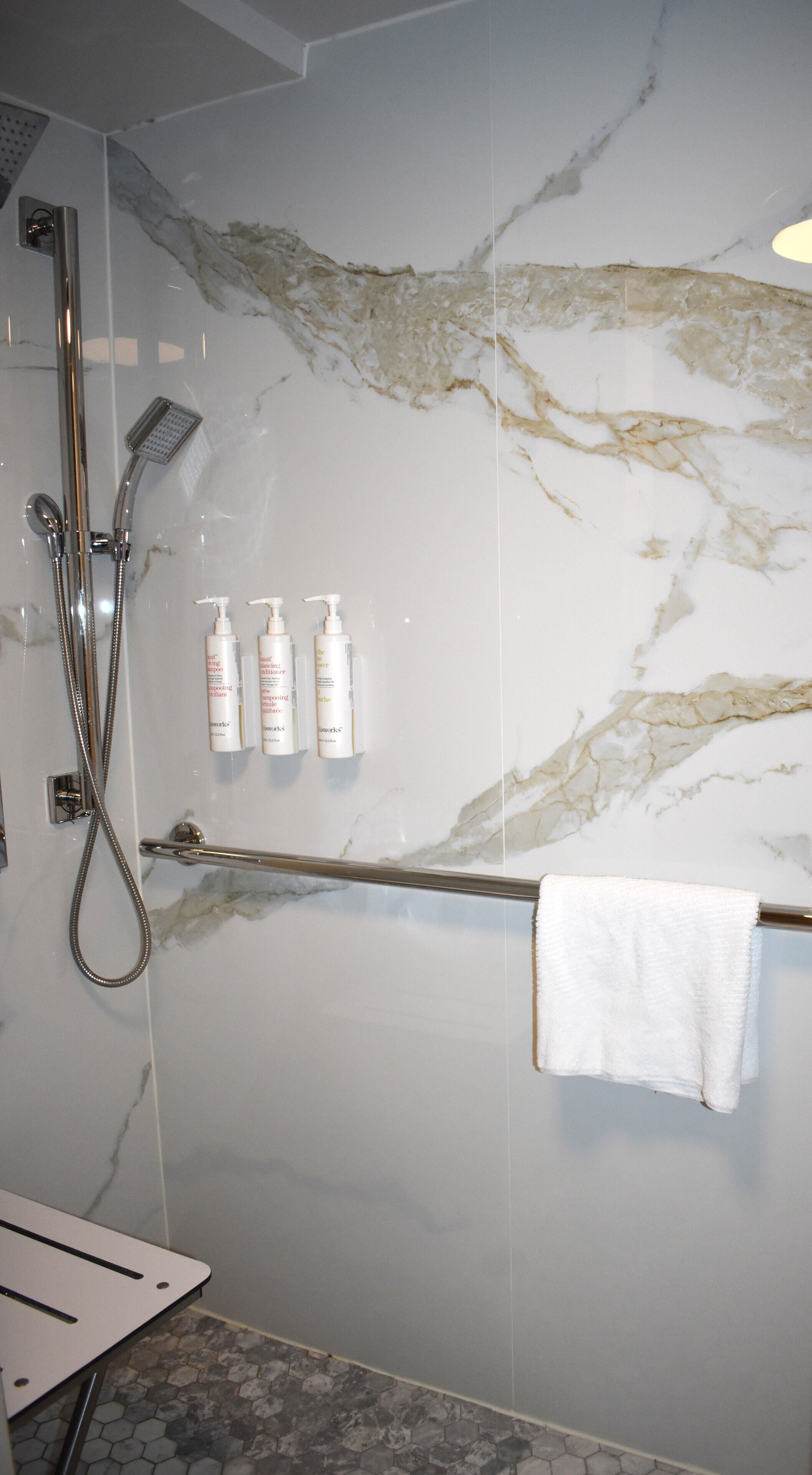 A bathroom with a marble wall, a shower with a handheld showerhead, a towel rack with a white towel, and two bottles of shampoo and conditioner.