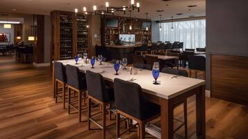Un restaurante con una larga mesa dispuesta para comer, una zona de bar con una estantería para vinos y vistas a la ciudad a través de grandes ventanales.