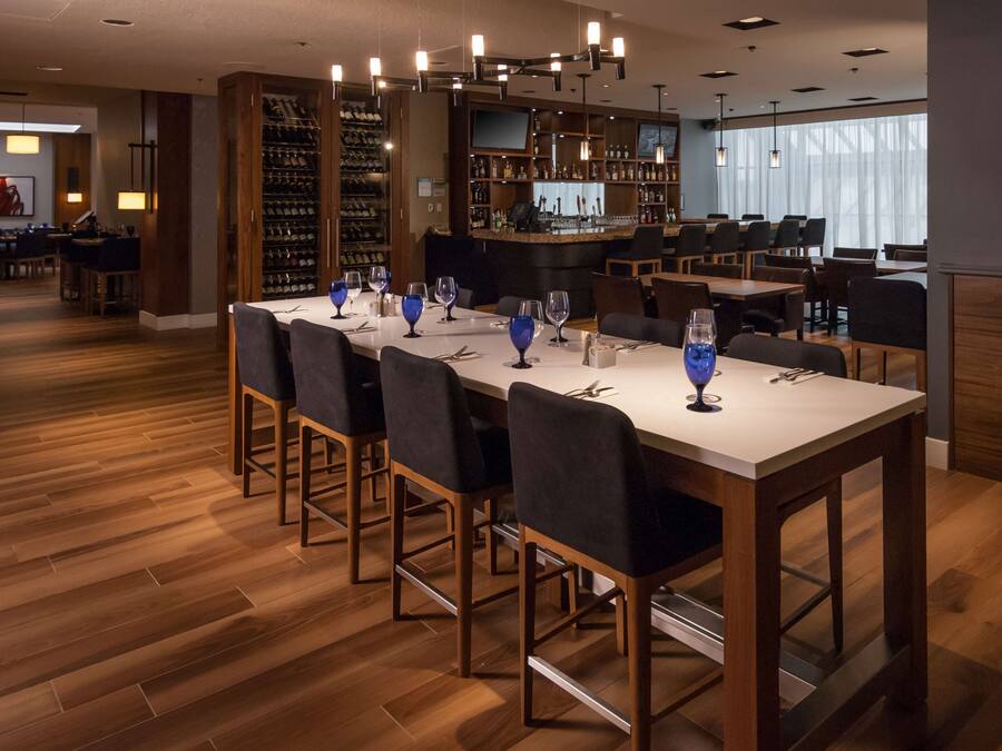 Un restaurante con una larga mesa dispuesta para comer, una zona de bar con una estantería para vinos y vistas a la ciudad a través de grandes ventanales.