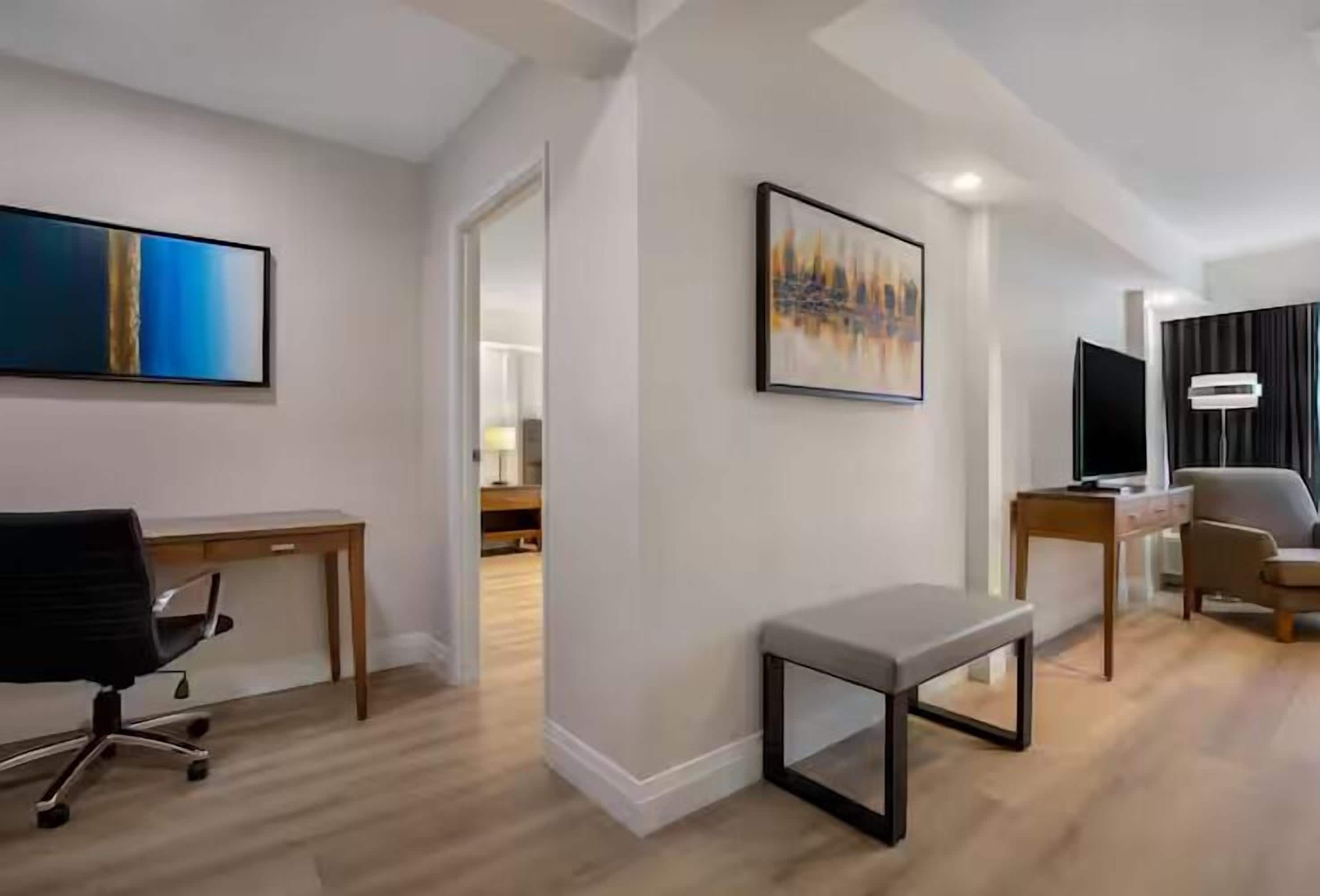 A modern hotel room with a wooden desk, a black office chair, a grey bench, and two framed artworks on the walls.