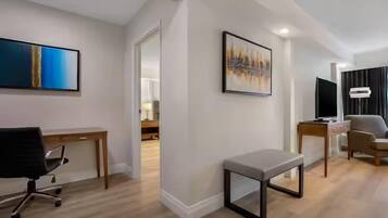 A modern hotel room with a wooden desk, a black office chair, a grey bench, and two framed artworks on the walls.