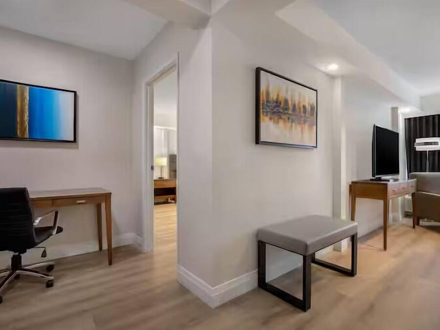 A modern hotel room with a wooden desk, a black office chair, a grey bench, and two framed artworks on the walls.