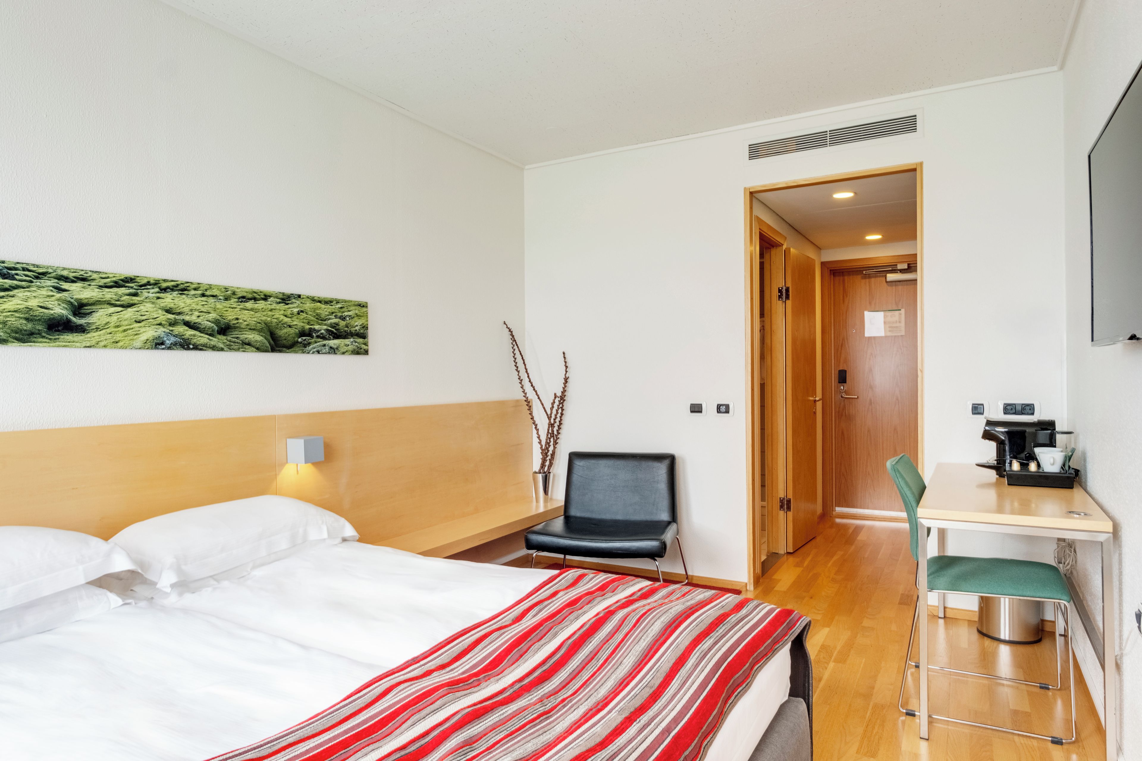 A hotel room with a bed, a desk with a chair, a coffee machine, and a door leading to another room.