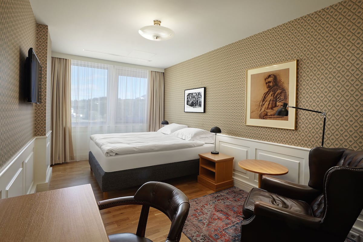 Une chambre d’hôtel avec un lit, un bureau, une chaise, une petite table, une lampe et un tableau au mur.