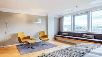 Habitación de hotel moderna con suelo de madera, sofá cama, dos sillones, una mesita y un ventanal con vistas a la ciudad.