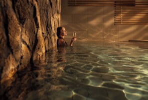 Une personne se trouve dans une piscine intérieure dont les murs sont en pierre, l’eau est claire, et elle tient un verre à la main.