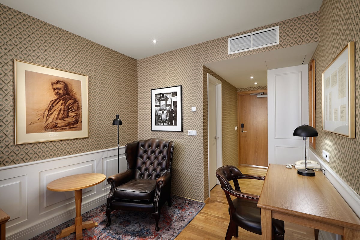 Une pièce avec un papier peint à motifs, un fauteuil en cuir capitonné, une table ronde en bois et un bureau avec une lampe.