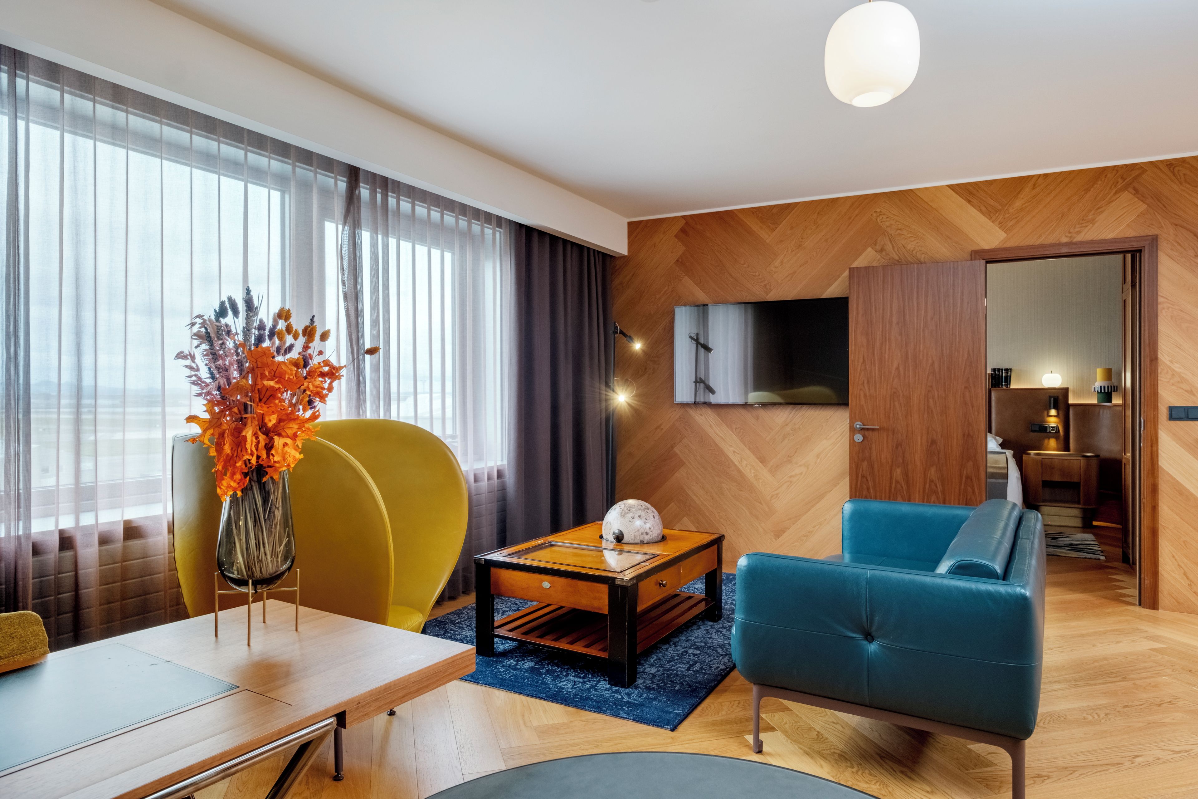 A modern living room with a wooden herringbone floor, a blue sofa, a yellow armchair, a wooden coffee table, and a glass vase with orange leaves.