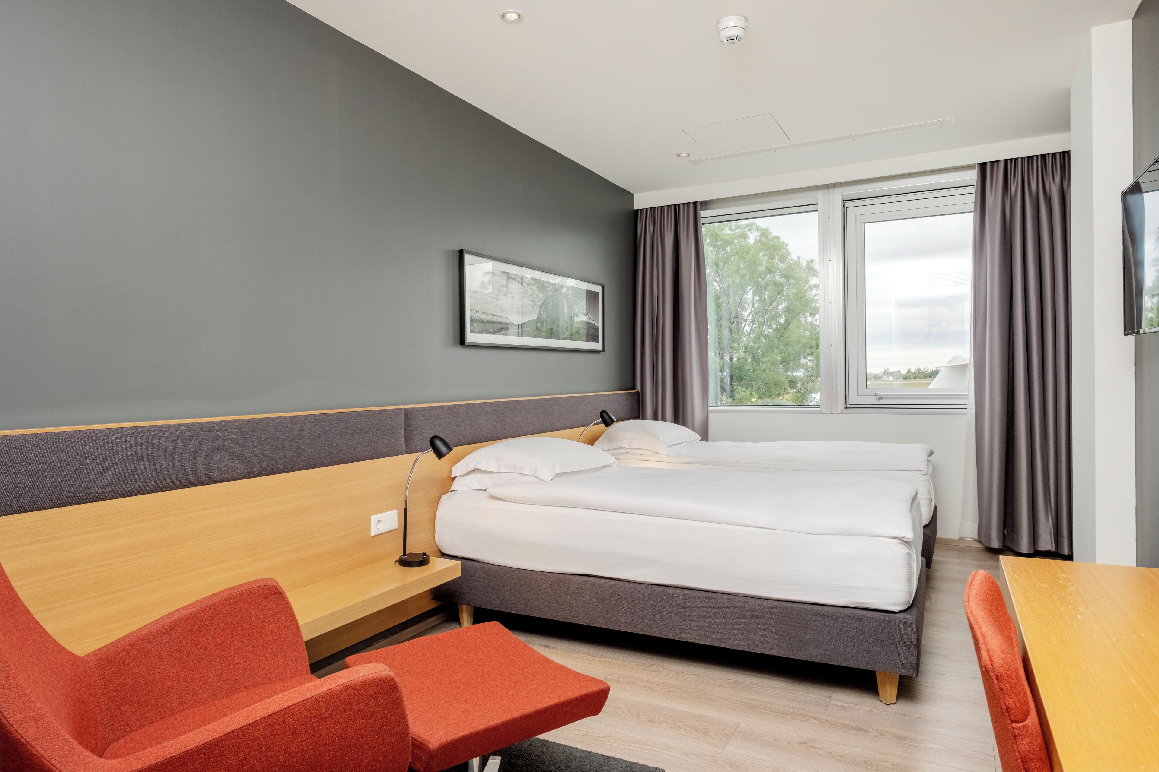 A hotel room with a bed, a desk, a chair, a bedside table, a lamp, a window with curtains, and a framed picture on the wall.