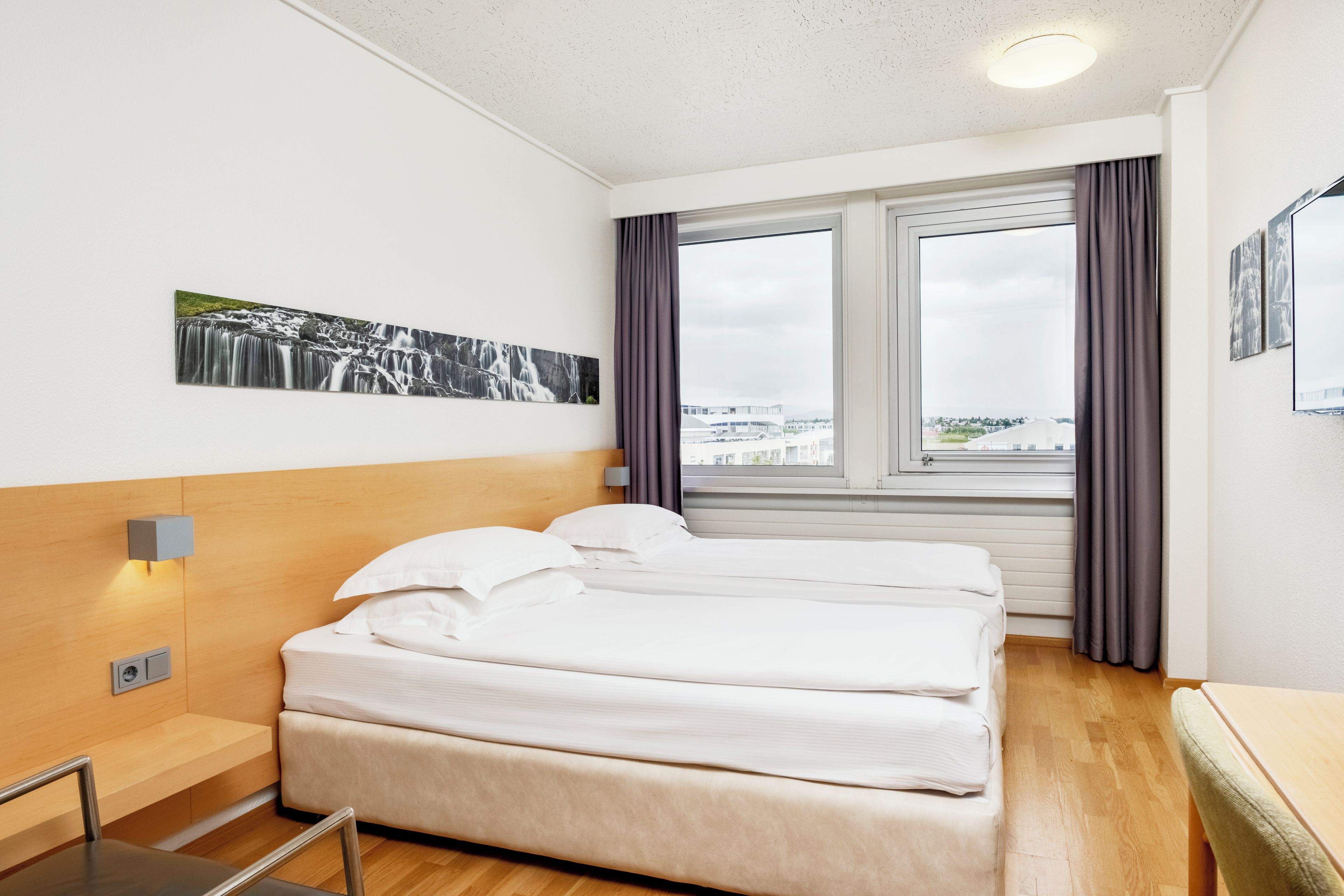 Habitación de hotel con cama, mesita de noche, lámpara de pared, ventana con cortinas y vista a la ciudad.