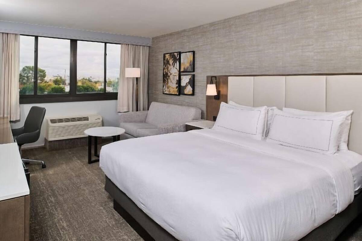 Habitación de hotel con una cama grande, un escritorio, una silla, una mesita y una ventana con cortinas.