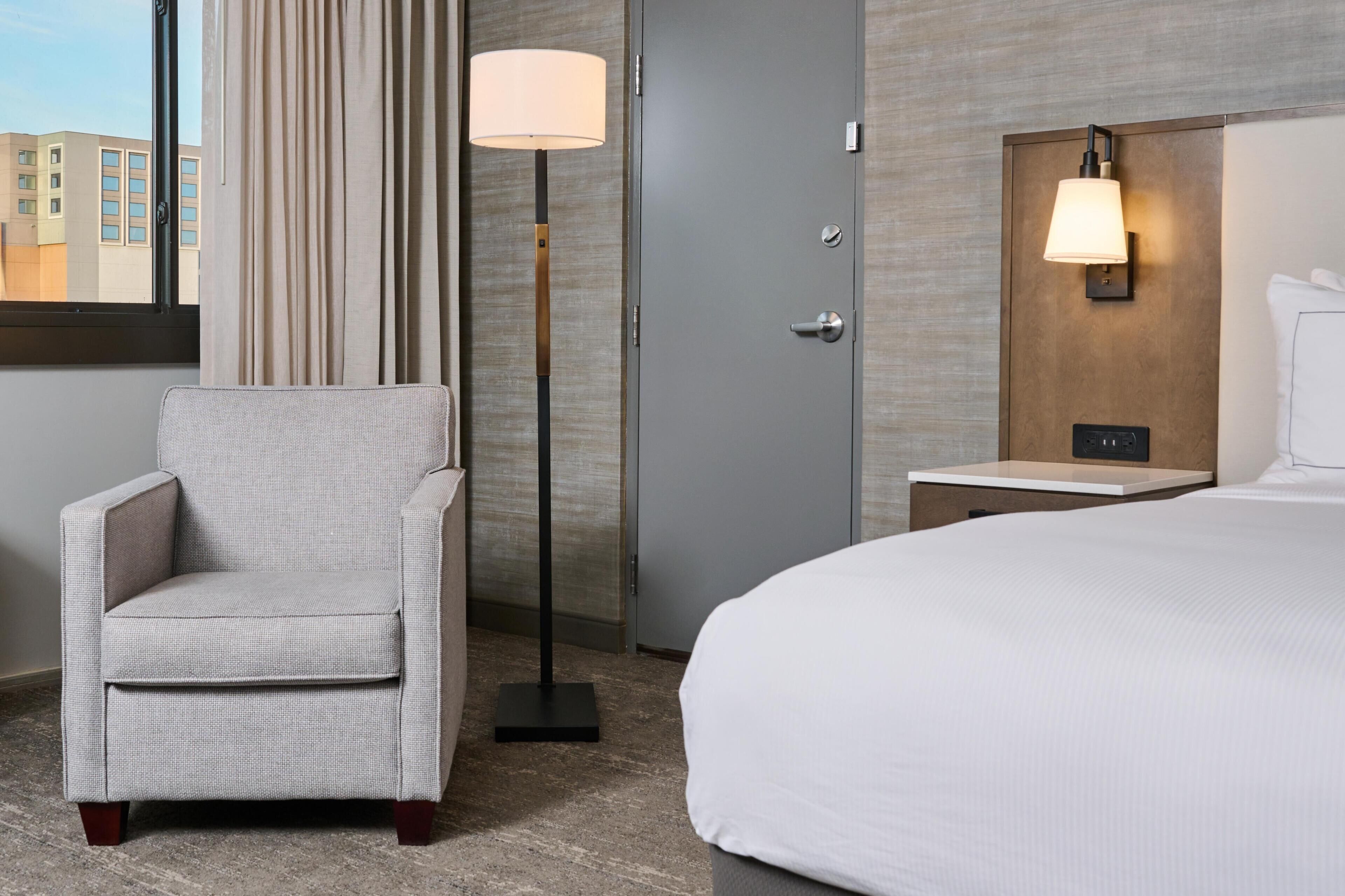 Una habitación de hotel con cama, una silla, una lámpara de pie, una mesita de noche y una lámpara fijada en la pared.