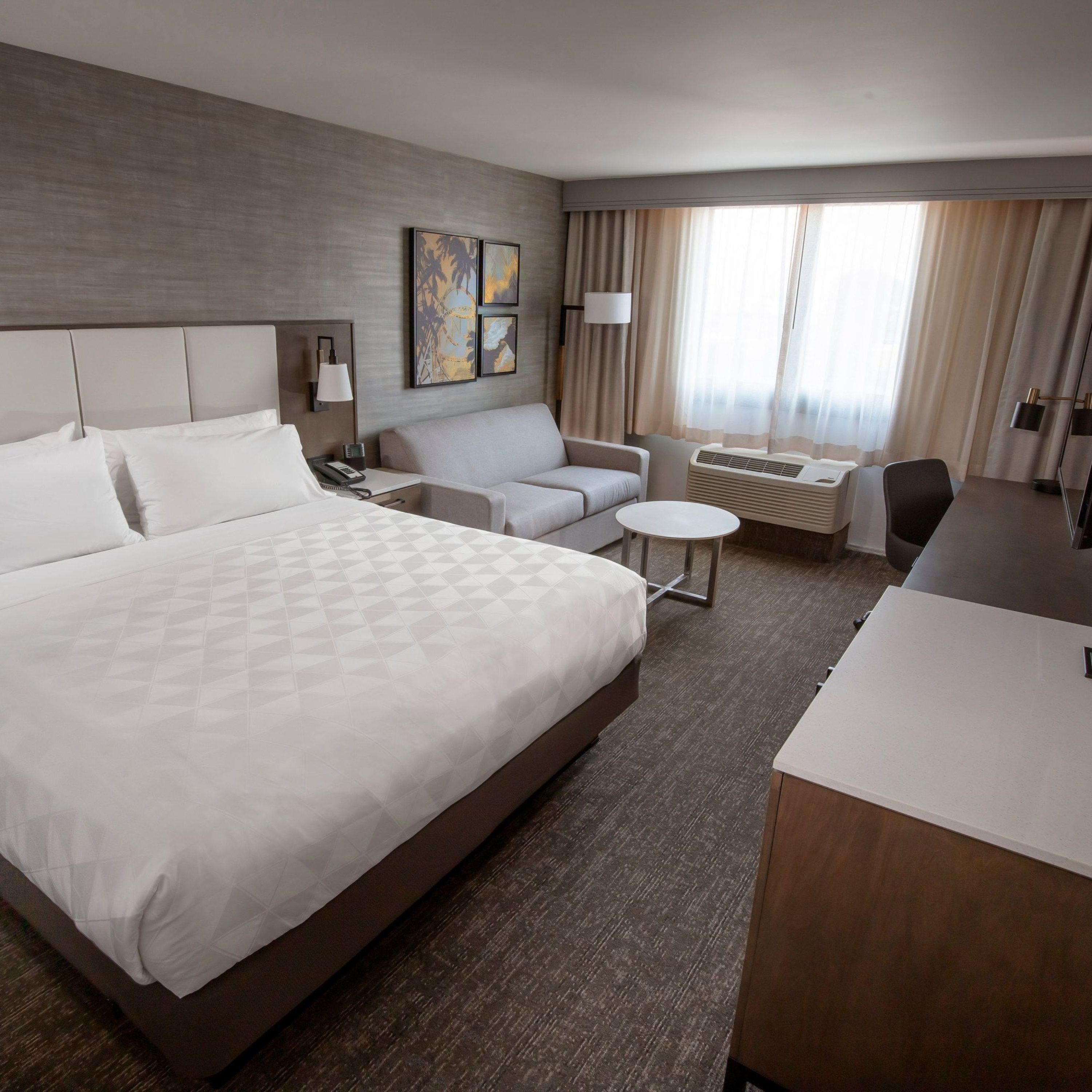 A hotel room with a large bed, a sofa, a desk, and a chair. There is a lamp, a phone, and a small table.