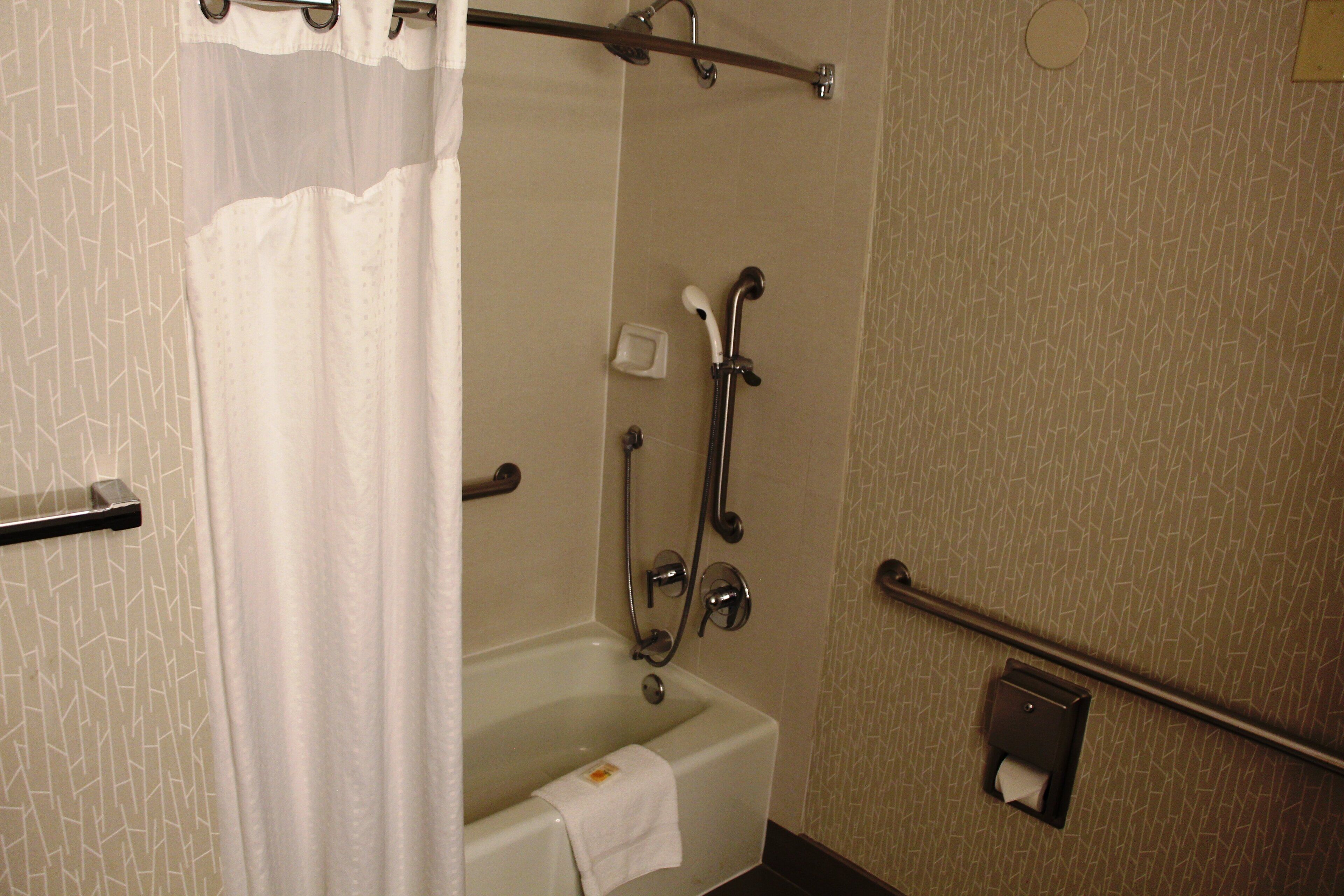 A bathroom with a bathtub, shower, handrails, a towel rack, and a soap dispenser.
