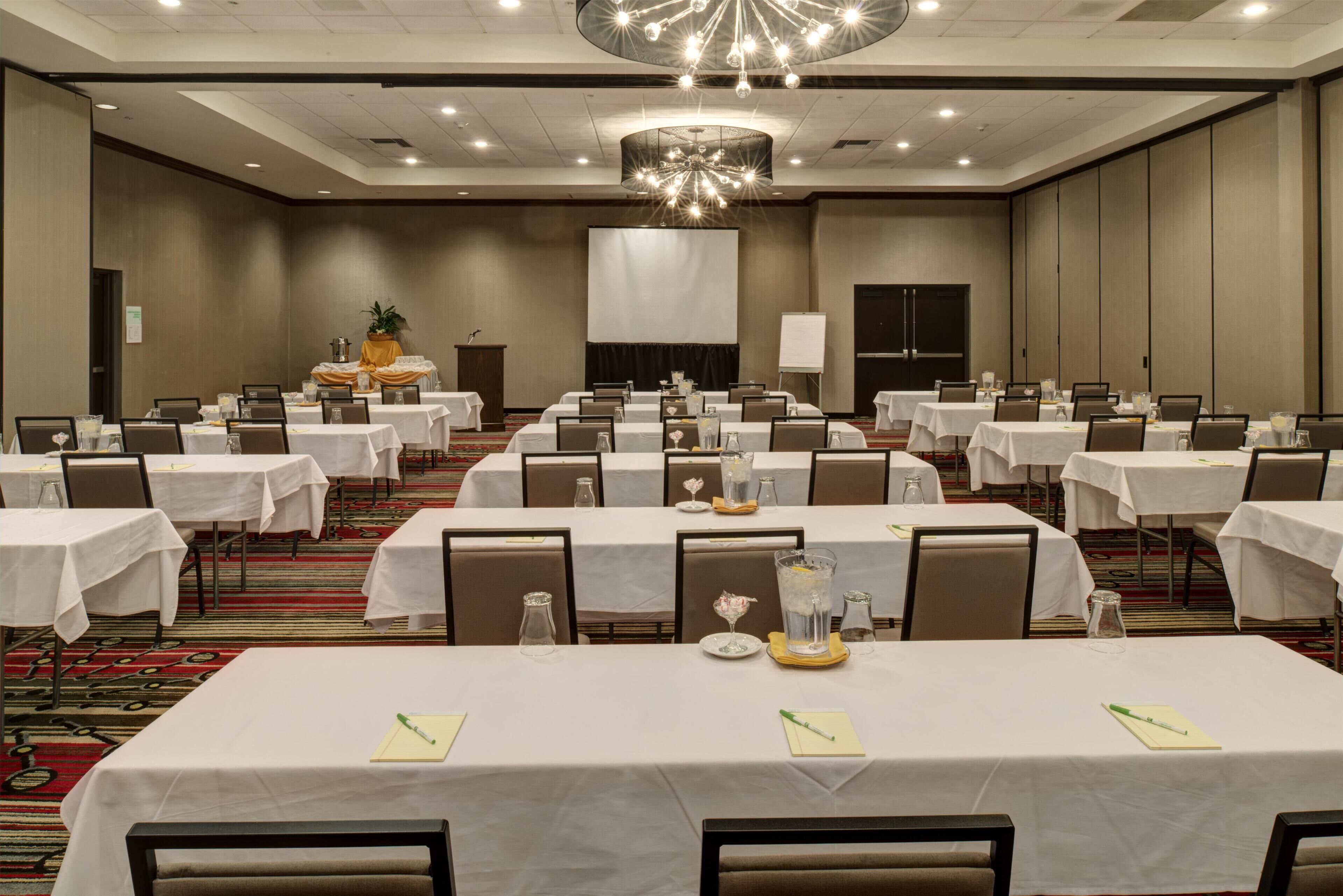 A conference room with multiple tables, chairs, a podium, and a large screen.