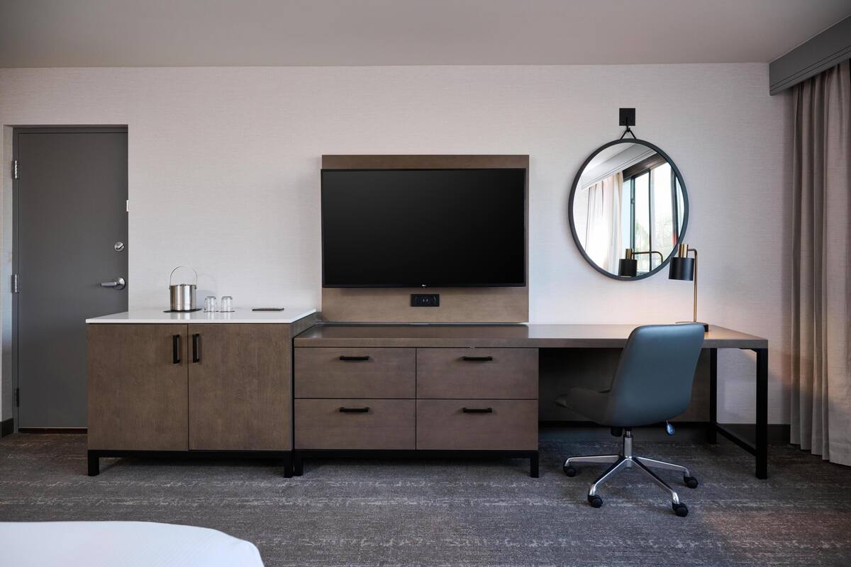 Una habitación de hotel con una cama, un escritorio con televisor, un espejo y una puerta.