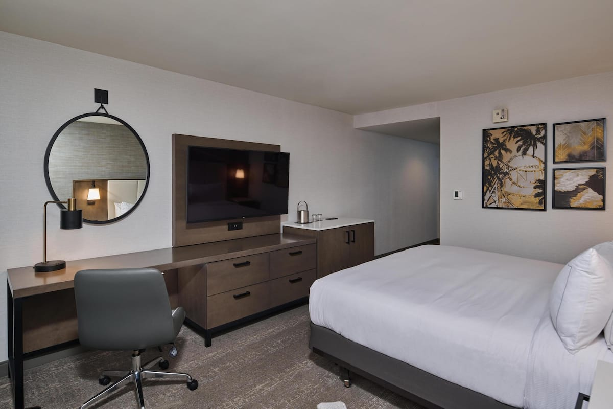 Habitación de hotel con cama, escritorio con silla, televisor, espejo y obras de arte en la pared.