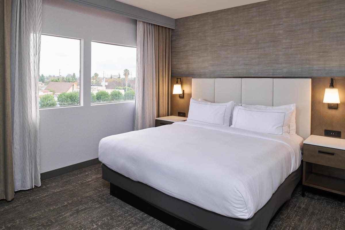 Habitación de hotel con una cama grande, dos mesitas de noche, un cabecero y un ventanal con cortinas.