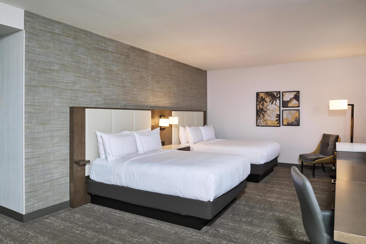 Habitación de hotel con dos camas, un escritorio, una silla y obras de arte en la pared.