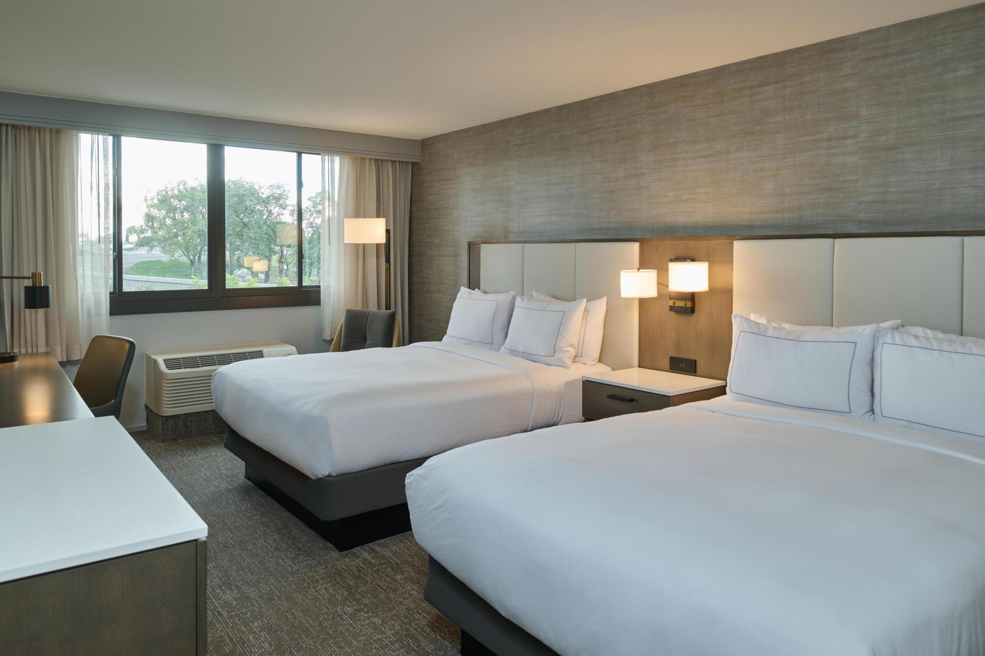 A hotel room with two beds, a desk, a chair, a window with curtains, and a wall-mounted lamp.