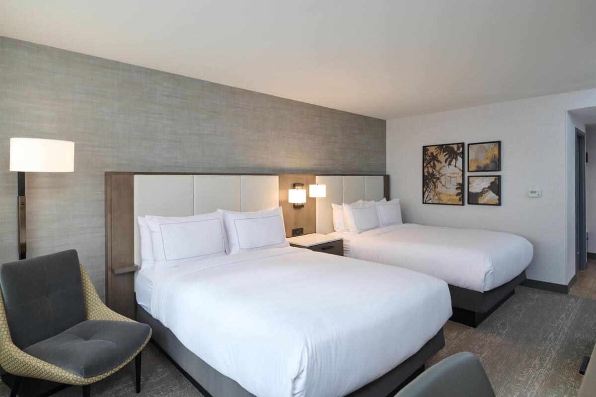 Una habitación de hotel con dos camas, una silla, una lámpara y una obra de arte en la pared.