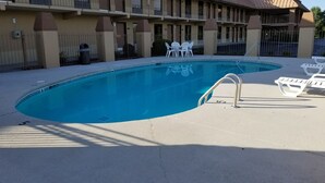A swimming pool with a curved edge, surrounded by a concrete deck. There are white lounge chairs and a small table with chairs.