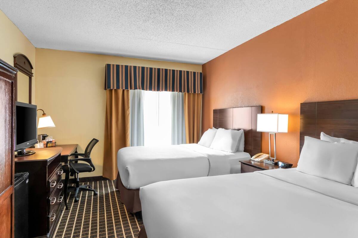 A hotel room with two beds, a desk with a TV, a chair, a dresser, and a window with curtains.