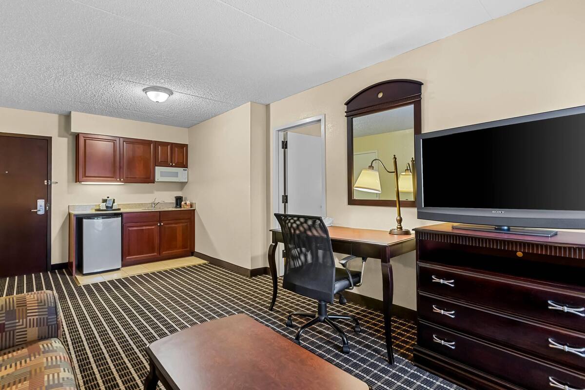 A hotel room with a kitchenette, a flat-screen TV, a desk, a chair, a dresser, and a patterned carpet.