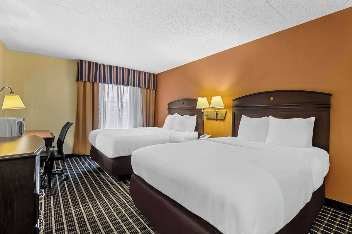 A hotel room with two beds, a desk with a chair, a lamp, and a window with curtains.