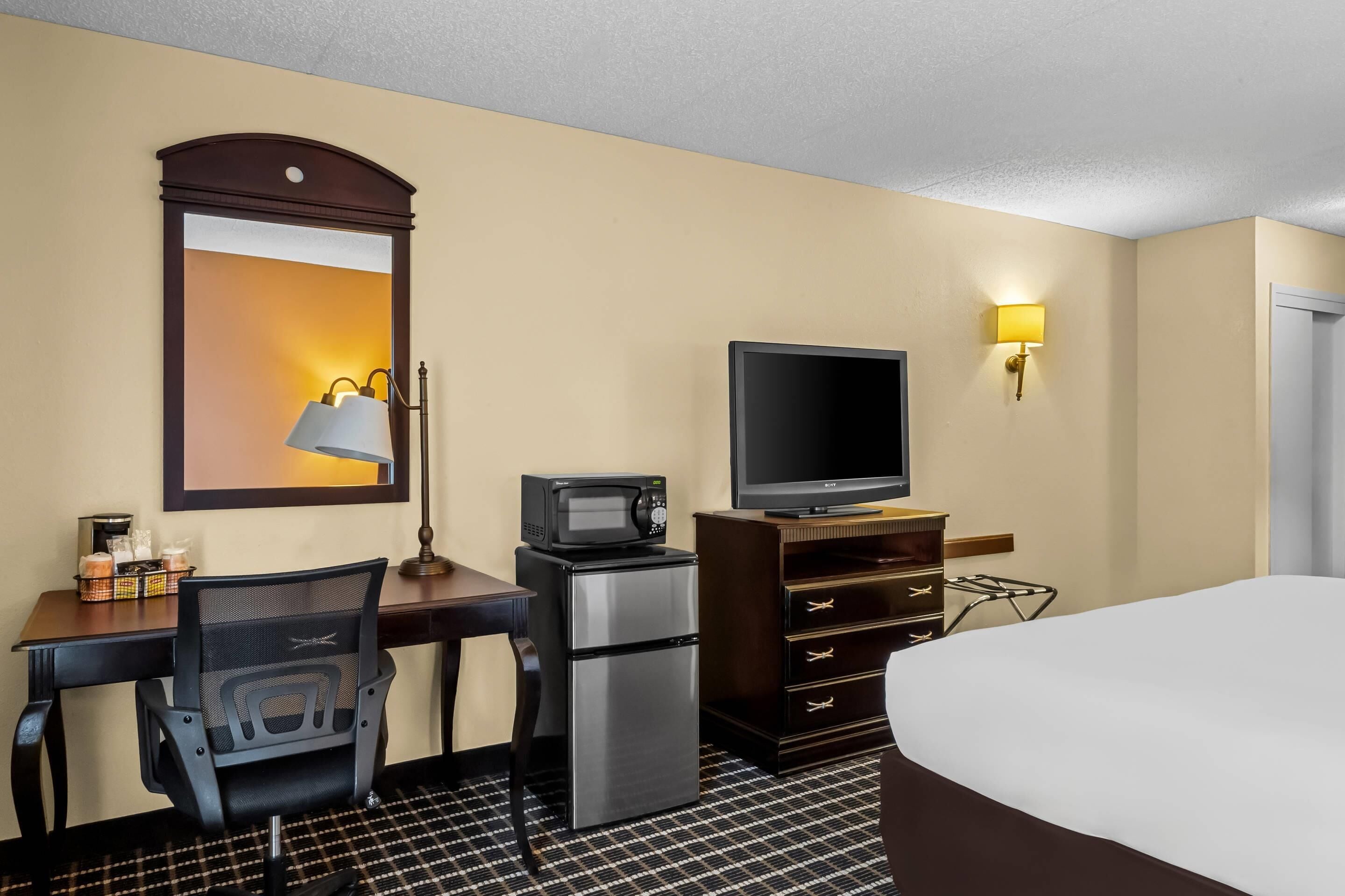 Habitación de hotel con cama, escritorio con silla, microondas, refrigerador, televisor, lámpara y luz empotrada en la pared.