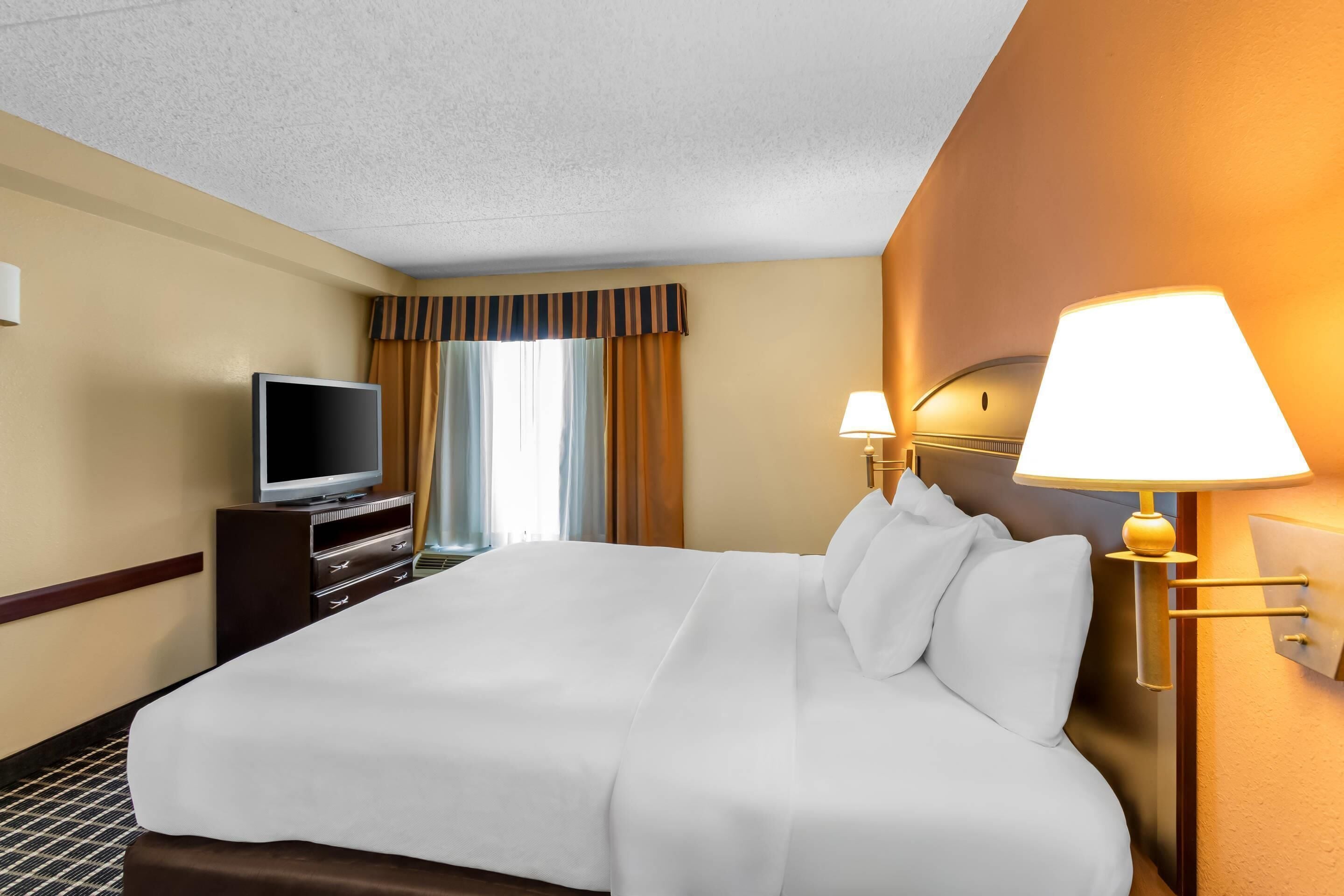 A hotel room with a large bed, a television on a dresser, two wall-mounted lamps, and a window with curtains.