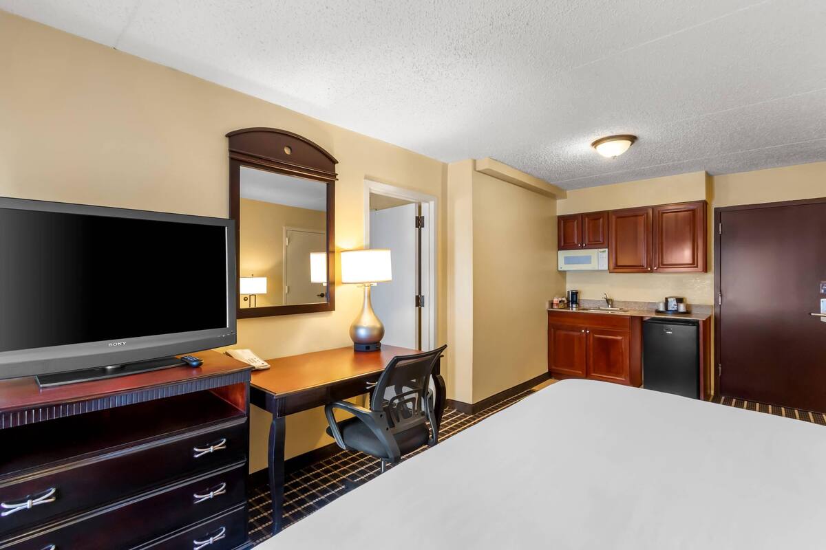 A hotel room with a bed, a TV, a desk with a chair, a mirror, a lamp, a microwave, and a refrigerator.