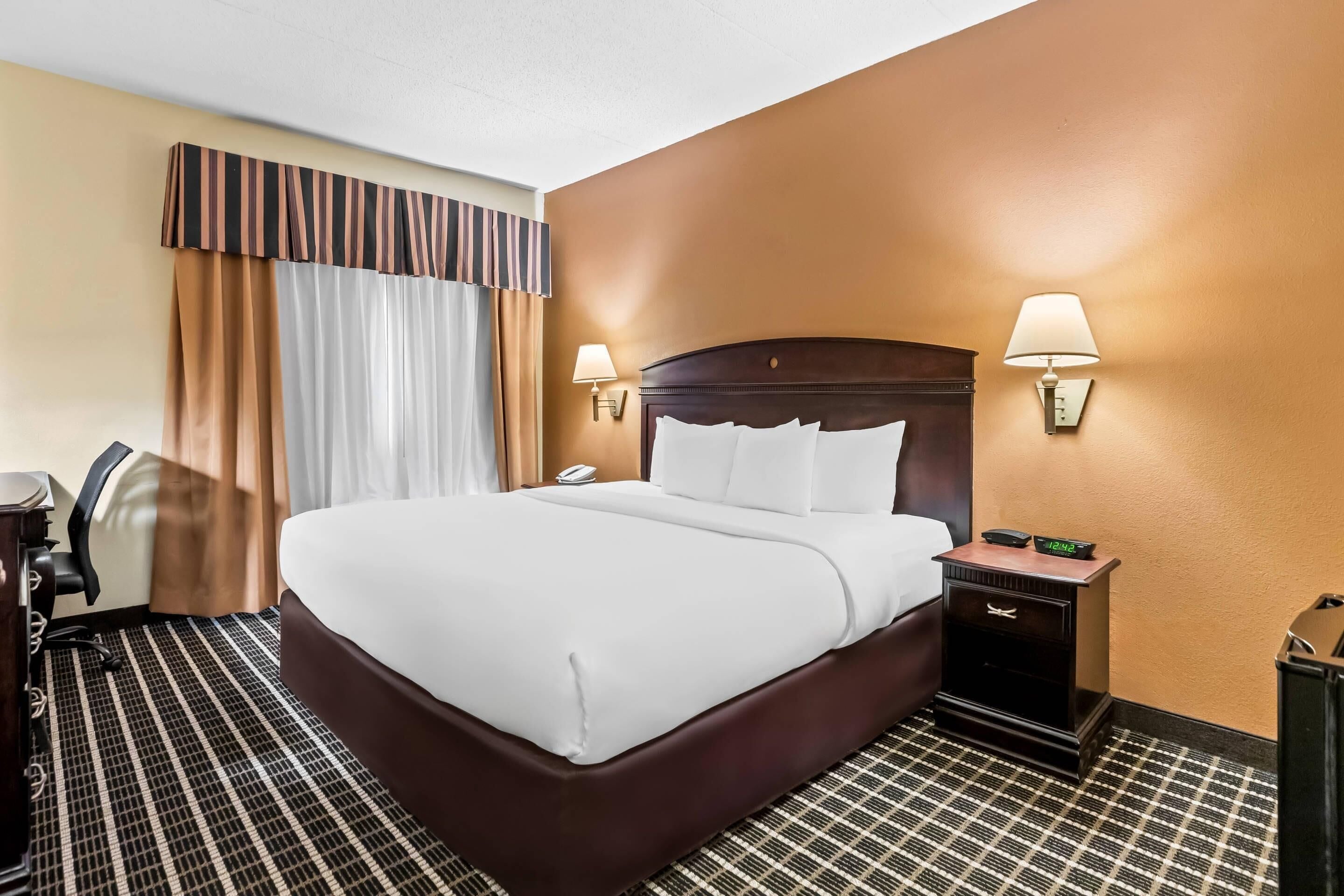 A hotel room with a large bed, two bedside lamps, a nightstand with a clock, a desk with a chair, and a window with curtains.