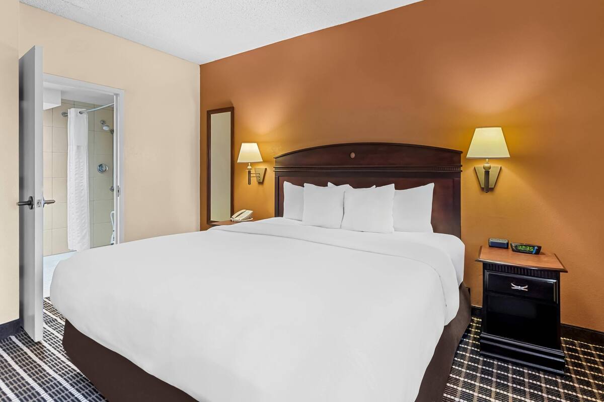 A hotel room with a large bed, two bedside lamps, a nightstand with a clock, and a door leading to a bathroom.