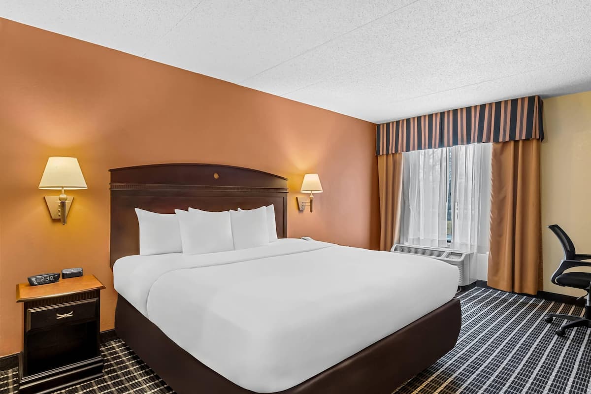 A hotel room with a large bed, two bedside lamps, a nightstand with a clock, a chair, and a window with curtains.