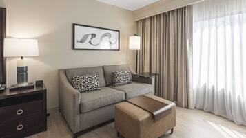 Una habitación de hotel moderna con un sofá gris, un reposapiés marrón, una obra de arte enmarcada en la pared y un ventanal con cortinas.