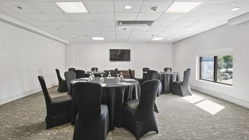 A conference room with round tables and black chairs, equipped with glassware and papers, featuring a large window and a flat-screen TV on the wall.