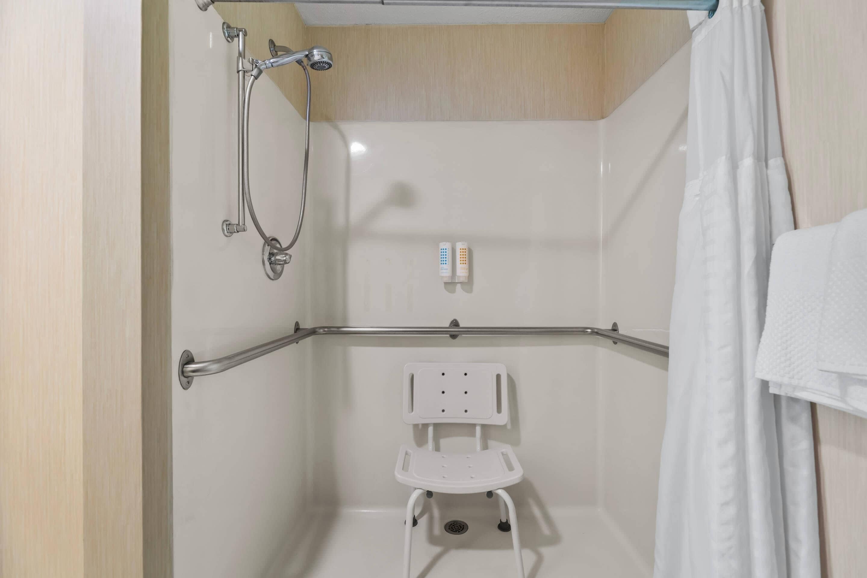 Zona de ducha con barra de apoyo, una silla blanca y ducha.