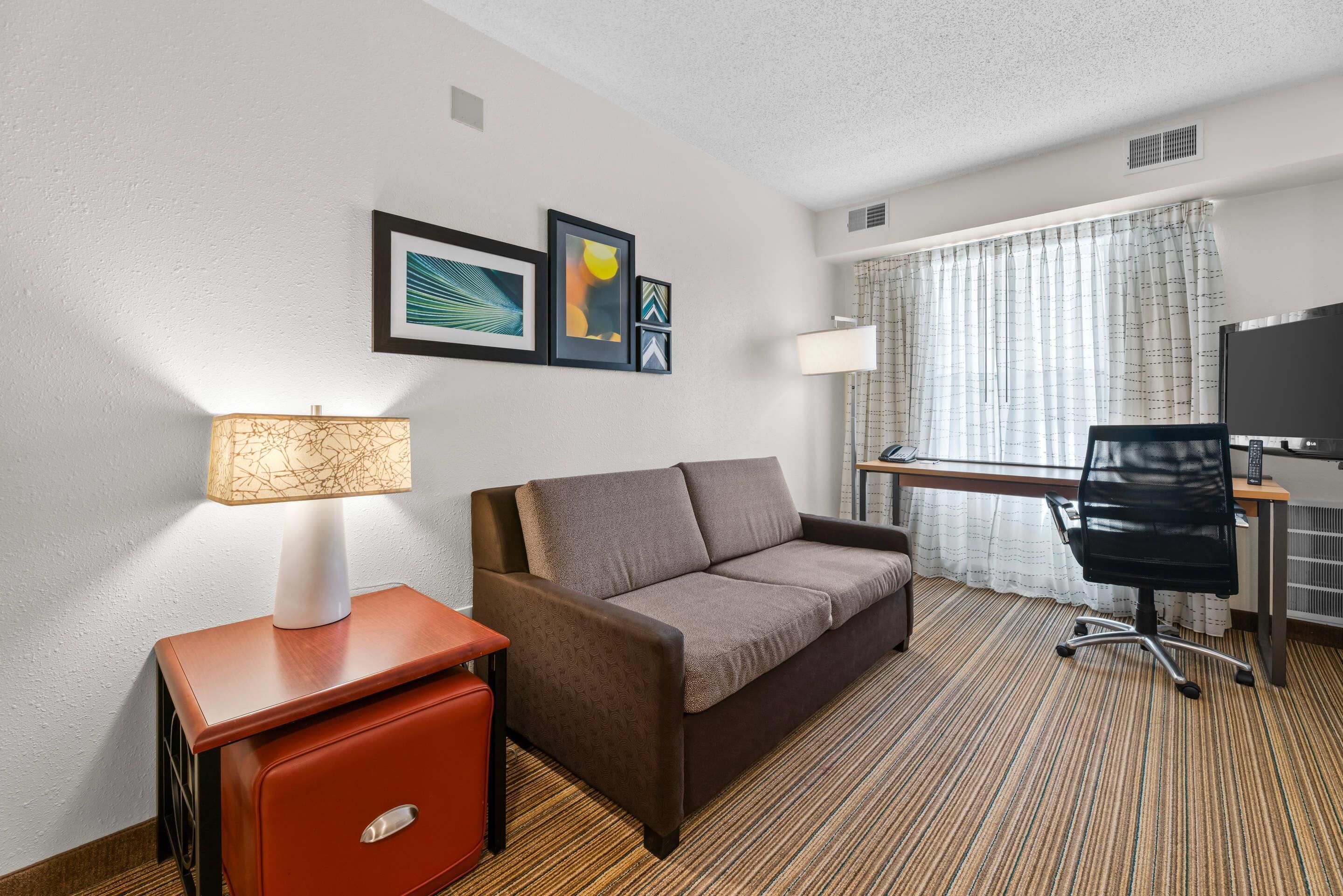 Une chambre d’hôtel avec un canapé, une petite table avec une lampe, un bureau avec une chaise et des œuvres d’art encadrées accrochées au mur.