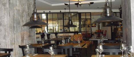 A restaurant interior with wooden tables, metal pendant lights, and a view of the kitchen through large windows.
