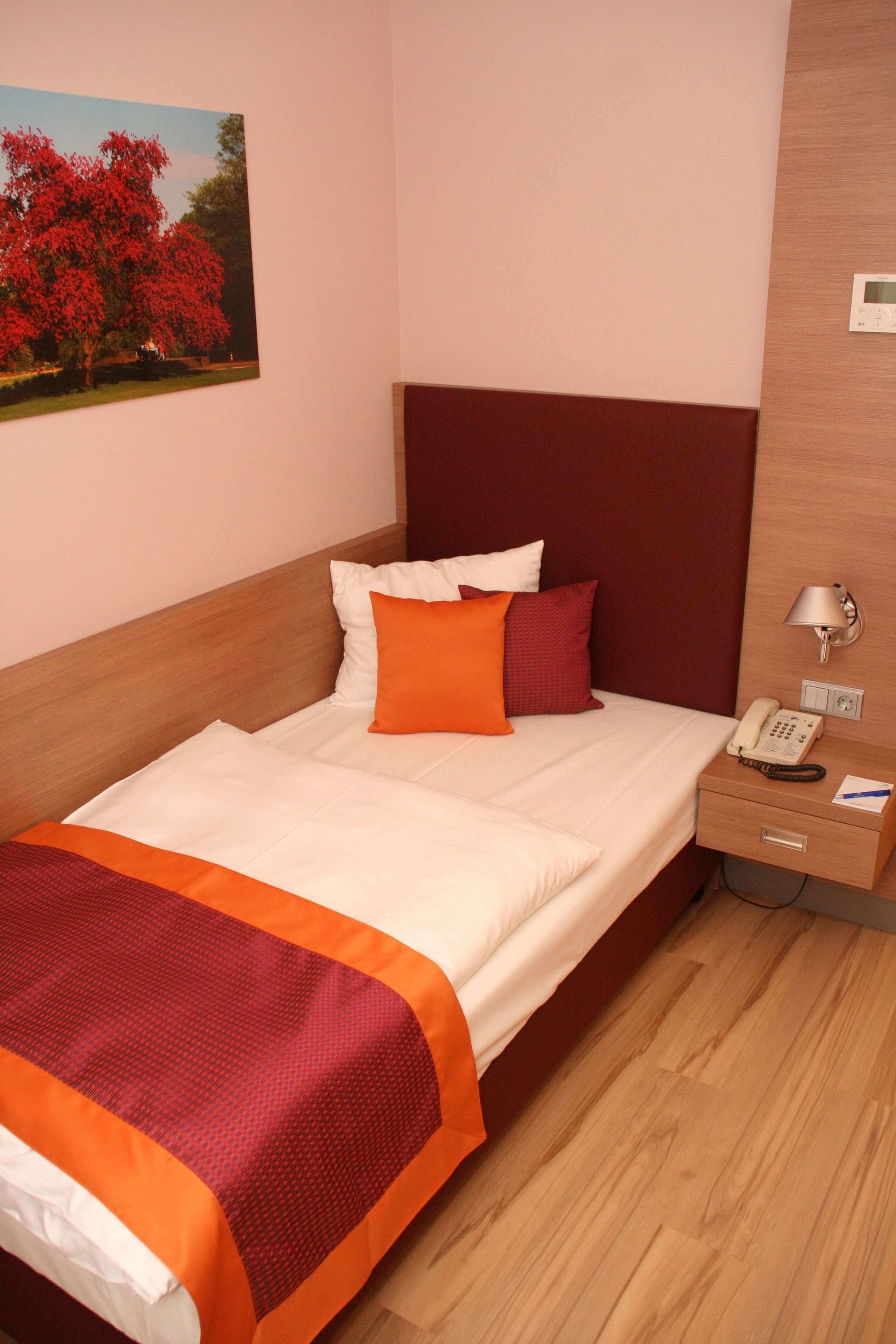 Una cama bien hecha con una colcha roja y blanca, un cabecero de madera y una mesita de noche con lámpara y teléfono.